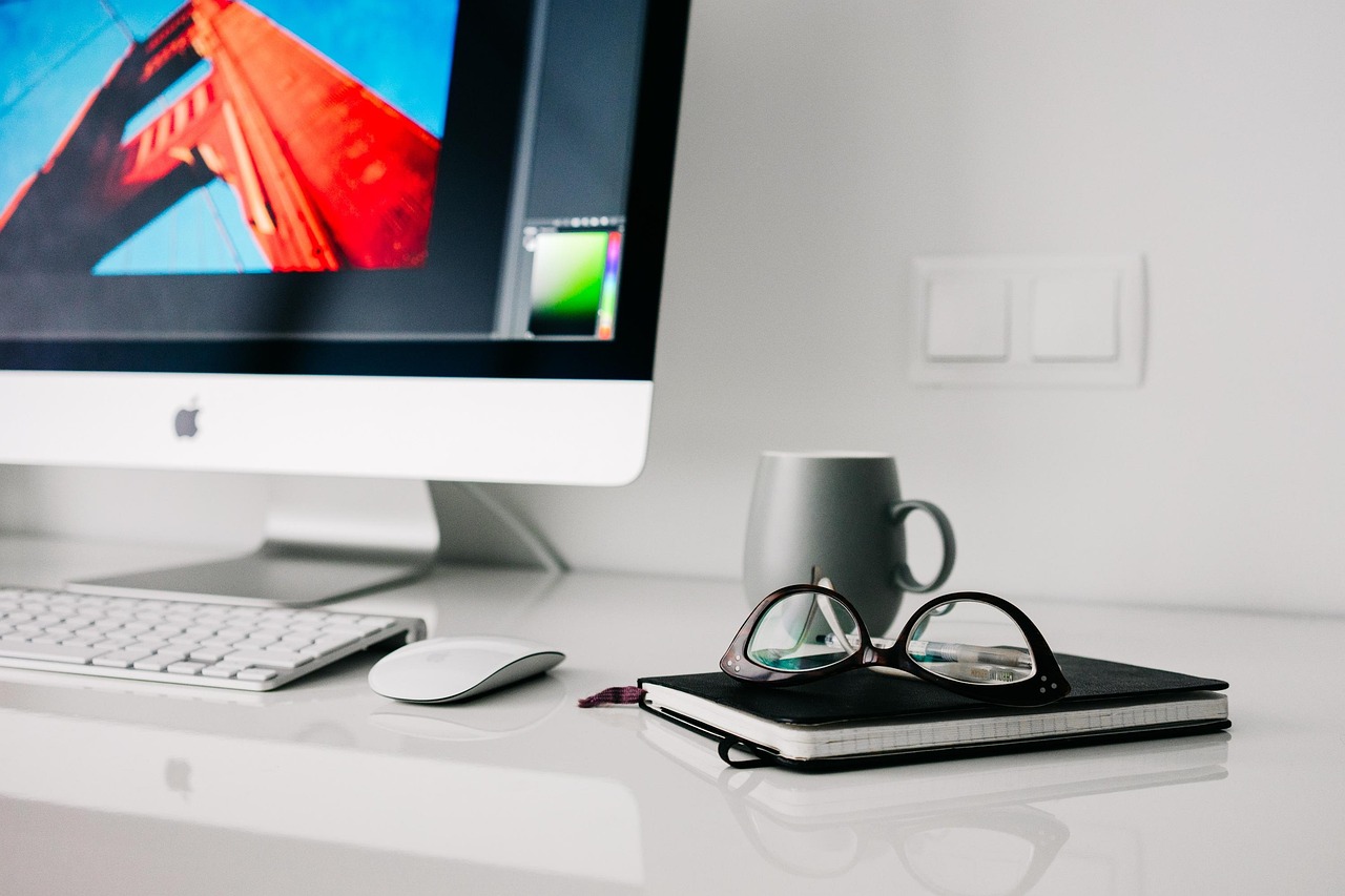 An iMac, a keyboard, a mouse, a notebook, a cup, and a pair of glasses on a white desk.